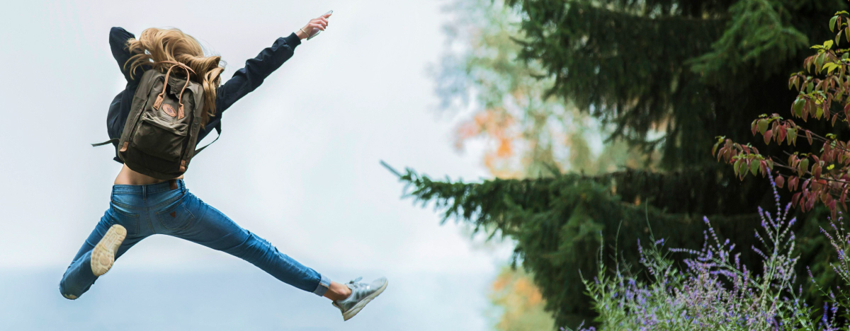 Eine Junge Frau mit Rucksack springt fröhlich durch den Wald.