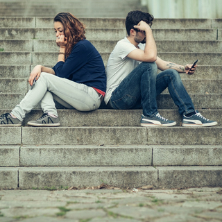 Eine Frau und ein Mann sitzen Rücken an Rücken auf einer Treppe und beachten sich nicht.