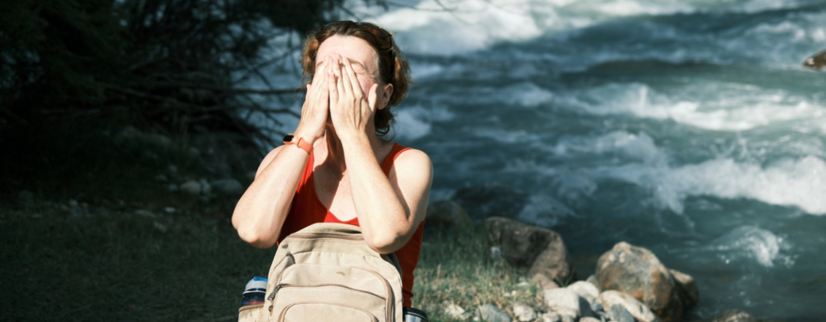 Frau steht in der Sonne an einem Fluss und hält ihre Hände über das Gesicht