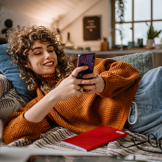 Junge Frau liegt auf ihrer Couch, während sie ihr mobiles Smartphone benutzt