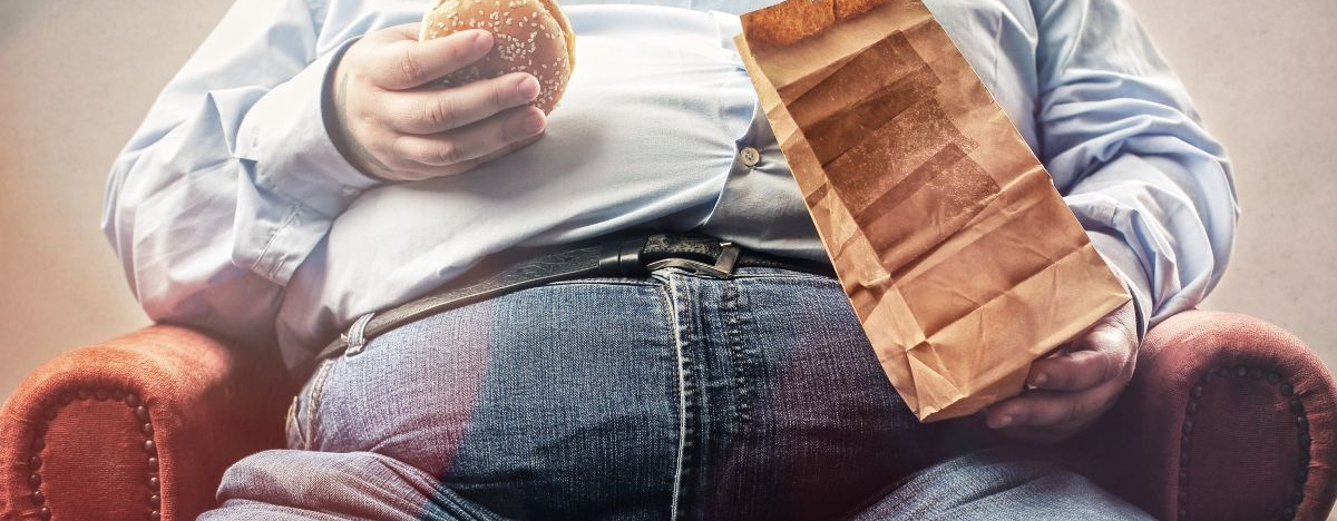 Dicker Mann sitzt auf dem Sofa und hält einen Burger in der Hand.