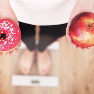 Frau steht auf Personenwaage und hält in der einen Hand einen Apfel, in der anderen einen Donut