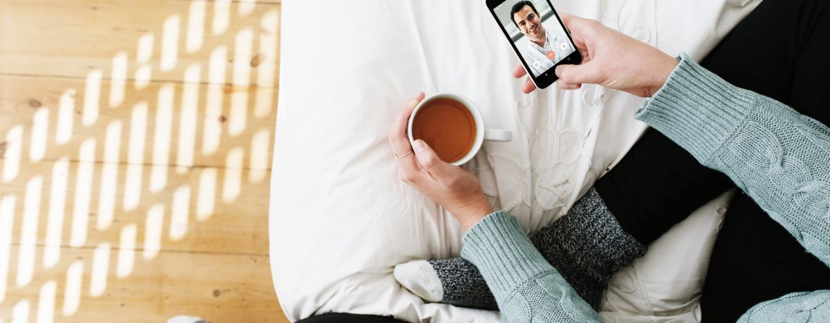 Eine Frau sitzt zuhause mit einer Tasse Kaffee und nutzt das Smartphone für eine Onlinesprechstunde der TeleClinik