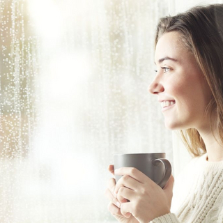 Frau steht lächelnd am Fenster und draußen regnet es