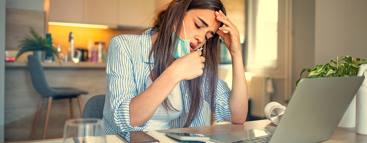 Eine Frau sitzt im Homeoffice, sie zieht sich die Maske vom Mund und hält sich den Kopf vor Schmerzen