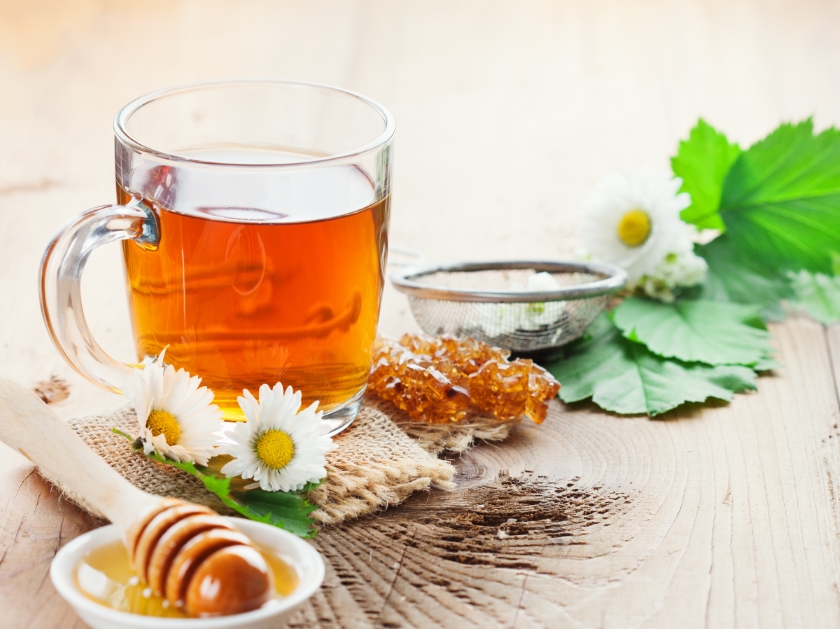 Ein kleines Glas mit Honig-Tee steht auf einem Holzuntergrund. Daneben liegen Gänseblümchen und ein Holzstab zum Honig entnehmen.
